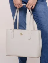 Cartera tipo tote de diseño semi-rígido y color beige, confeccionada en eco cuero. Cuenta con doble manija de hombro, base reforzada con remaches metálicos y aplique dorado con el logo de la marca en el frente.