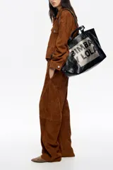 Bolso tipo tote de Bimba y Lola, color dorado con acabado metalizado y efecto arrugado. Presenta un diseño cuadrado con estampado central de la marca en un recuadro oscuro y doble asa de mano en color crema.