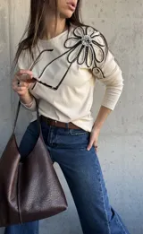 Remera manga larga de color beige, con detalle de aplique floral tridimensional en el hombro confeccionado con cordones en blanco y negro.