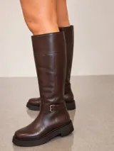 Bota de caña alta confeccionada en cuero color marrón chocolate, con diseño de plataforma y taco bajo. Presenta una tira decorativa con hebilla metálica en la zona del tobillo y una costura horizontal en la parte superior de la caña.