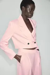 Blazer corto rosado con cuello solapa, hombreras y mangas largas. Cuenta con cierre cruzado delantero con dos botones oscuros y un bolsillo de ojal en el pecho.