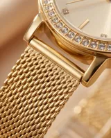 Reloj analógico de mujer con caja redonda dorada, bisel con incrustaciones de cristales y malla de acero tipo milanesa. Esfera minimalista con índices puntuales.