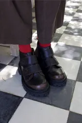 Borcego de cuero color marrón oscuro con acabado desgastado en la puntera. Presenta un diseño de caña corta con cierre lateral y dos tiras ajustables con velcro en el empeine. Cuenta con una suela de goma robusta tipo tractor.