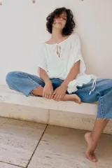 Top blanco de cuello redondo y mangas tipo capa, con textura fruncida, bajo asimétrico, lazos decorativos en el escote y en los laterales, y cierre posterior con botón.