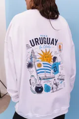 Buzo oversize de cuello redondo en color blanco, con estampa gráfica en la espalda que incluye motivos uruguayos, el sol, la bandera, un jugador de fútbol y elementos tradicionales.