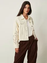 Campera de denim color off white con cuello camisero, bolsillos con solapa en el pecho y cierre frontal con botones metálicos. Presenta detalles de bordado con perforaciones en las mangas.