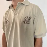 Chomba de corte oversize color beige, con cuello polo y cierre frontal de botones. Presenta bordados en el pecho con motivos de golf y tipografía cursiva.
