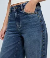 Pantalón wide leg de jeans azul oscuro, con cintura media y detalle de tachuelas metálicas a lo largo de las costuras laterales. Cuenta con bolsillos funcionales, trabillas para cinturón y cierre frontal con botón y cremallera.