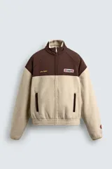 Cazadora de corte relajado confeccionada en tejido de borreguillo color beige combinado con tejido técnico marrón en la parte superior. Presenta cuello subido, cierre frontal con cremallera, bolsillos laterales de vivo y puños y bajo elásticos. Incluye parches y bordados decorativos en el pecho.