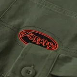 Camisa de manga larga color verde militar, con cuello clásico, cierre de botones frontales, dos bolsillos en el pecho y un parche ovalado con texto en rojo en el bolsillo izquierdo.