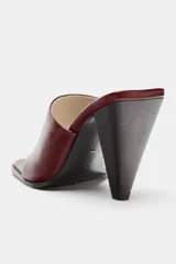 Sandalia de tacón en piel color bordó, con diseño tipo mule y tira delantera con separación para el dedo. Cuenta con tacón alto asimétrico y punta cuadrada.
