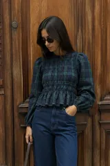 Blusa de manga larga con diseño de cuadros escoceses en tonos verde y azul. Presenta un cuerpo con frunces elásticos que crean un efecto ajustado y un pequeño volado en el ruedo. Las mangas son abullonadas con puños elásticos.