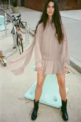 Vestido corto de tejido fluido en color beige rosado, con cuello subido ajustable mediante lazada en la espalda. Presenta mangas largas abullonadas con puños abotonados, cintura entallada y falda con bajo acabado en volantes. Incluye forro interior y cierre lateral con cremallera oculta.