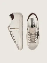 Zapatillas Golden Goose modelo Super-Star, color blanco con detalles en marrón con glitter. Confeccionadas en cuero vacuno, con estrella lateral y contrafuerte de talón brillantes, punta y lengüeta de gamuza, y efecto envejecido en la suela.