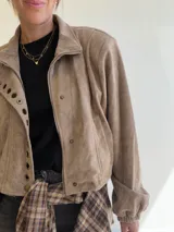 Chaqueta corta de efecto gamuza con cierre frontal y botones. Presenta mangas tipo globo con puños elásticos y detalle de aros metálicos decorativos en la solapa. Cuenta con ajuste lateral en la cintura y forro interior.