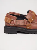 Mocasines Mutma Gales de cuero color tostado, con plataforma gruesa dentada, detalles de tachas metálicas, argollas y tira lateral con hebilla.