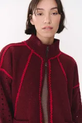 Campera de punto color bordó con destellos de lurex, diseño oversize, cuello alto y cierre frontal. Presenta detalles de costuras decorativas en color rojo vibrante y textura calada en las mangas.