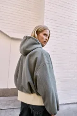 Chaqueta estilo bomber de corte oversize en color marrón oscuro, confeccionada en tejido suave. Presenta capucha con cordones ajustables, cierre frontal con cremallera, mangas largas con puños elásticos y bolsillos delanteros de vivo. El bajo cuenta con detalle de pliegues.