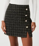 Pollera corta de tweed texturizado en color negro con hilos dorados, presenta botones decorativos con centro blanco y una pequeña abertura frontal en el ruedo.