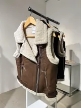 Chaleco estilo gamulán de cuero sintético marrón con interior y solapas de corderito sintético color crema. Presenta cierre frontal cruzado, bolsillos laterales con cierre y correas ajustables en los laterales del ruedo.