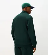 Buzo deportivo Get Over de color verde oscuro, confeccionado en tejido de algodón y poliéster. Presenta cuello alto, cierre frontal completo con cremallera, mangas largas con puños ajustados y bolsillos laterales. Incluye un pequeño lettering bordado en el pecho.