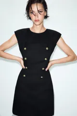Vestido corto de estilo militar en color negro, con cuello redondo, hombreras y manga sisa. Presenta un diseño frontal con falsos botones dorados dispuestos en filas horizontales. Cierre lateral con cremallera oculta y abertura en la espalda con botón.