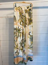 Pantalón palazzo de satén blanco con estampado barroco de hojas tropicales en tonos verde oliva y mostaza, y detalles de cadenas en color dorado y rosado. Presenta un borde inferior con detalles en tonos melocotón y azul claro.