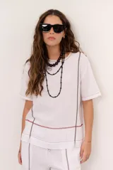 Blusa de lino color beige, de corte recto y mangas cortas, con costuras decorativas pespunteadas en tono rojizo que forman líneas verticales y horizontales.