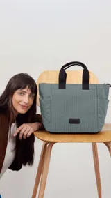 Cartera tipo tote de diseño capitoneado en color gris, con doble asa de mano en color negro y bolsillos laterales. Presenta un parche con logo en el frente y un diseño espacioso ideal para uso diario.
