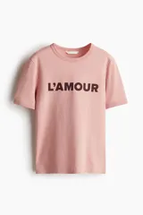 Remera de manga corta y cuello redondo color rosa pálido con la inscripción "L'AMOUR" en color marrón oscuro en el pecho.