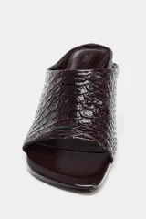 Sandalia tipo mule de tacón alto, color marrón oscuro con textura de piel de cocodrilo. Presenta punta cuadrada y un diseño abierto en la parte delantera.
