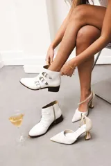 Botas cortas de cuero blanco hasta el tobillo, con puntera afilada, taco bajo y tres filas de correas con hebillas metálicas y tachas decorativas.