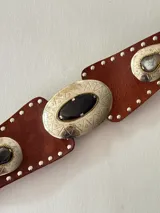 Cinto tipo faja de cuero color chocolate, con apliques metálicos plateados grabados y piedras incrustadas. Presenta detalles de tachas pequeñas en todo el borde y un diseño central ovalado.