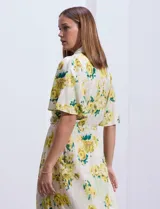 Vestido largo camisero con estampado floral amarillo y verde sobre fondo blanco. Tiene cuello camisero, mangas cortas con volados, cintura con lazo ajustable y falda escalonada con volados.