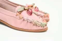 Mocasín plano de cuero color rosado, con punta redondeada y taco bajo. Presenta un detalle artesanal de tiras entrelazadas en tonos nude, verde y rosado alrededor del empeine, rematado con un aplique colgante central con piedra transparente.