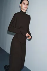 Vestido midi de punto color marrón, con cuello subido, manga larga y diseño cut out en un hombro. Presenta frunces laterales en la cintura que aportan un efecto drapeado.