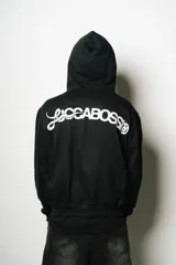 Hoodie con cierre frontal, color negro, de corte regular fit. Presenta un estampado frontal grande en blanco con un diseño abstracto y estilizado, similar a un monograma o símbolo.