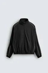 Chaqueta técnica de corte relajado en color negro, con cuello subido, cierre frontal de cremallera y detalles de líneas en contraste. Presenta bolsillos frontales con cierre oculto y acabados elásticos en puños y ruedo.