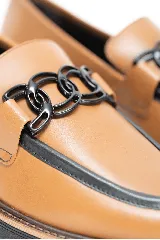Mocasín de cuero color marrón claro, con detalles en negro en el borde superior y suela plana del mismo color. Cuenta con un adorno de cadena plástica negra en el empeine.