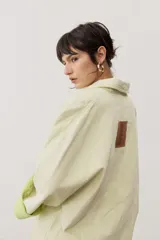 Chaqueta oversize de gabardina color verde claro, con solapa clásica, bolsillos frontales, cierre de botones oscuros y mangas largas con vuelta en tono verde lima vibrante.