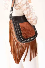 Cartera de hombro estilo western, confeccionada en una combinación de cuero vacuno negro, gamuza color óxido y cuero grabado color whisky. Presenta un diseño con flecos largos en el frente y los laterales, decorada con apliques metálicos tipo tachas a lo largo de la estructura. Incluye una correa corta fija y una correa larga extensible y desmontable.