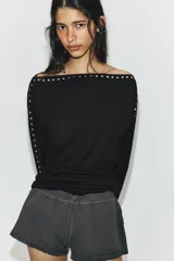 Camiseta de cuello barco y manga larga tipo murciélago, confeccionada en tejido de punto gris melange. Presenta detalle de tachas metálicas a lo largo de los hombros y las mangas.