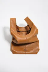 Bolso de mano Bimba y Lola modelo Paper mediano, confeccionado en cuero color tostado con estructura blanda, asas superiores y correa de hombro ajustable en color beige. Presenta costuras visibles, logo metálico de la marca en la parte inferior y cierre principal.