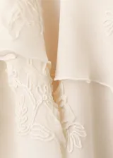 Blusa color marfil de tejido fluido, con cuello alto con lazo, volados frontales y mangas largas acampanadas con detalles florales bordados.
