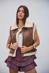 Chaleco de estilo aviador en color camel, confeccionado en material efecto cuero con interior y bordes de corderito sintético color crema. Presenta cierre frontal, cuello solapa y detalles de hebillas metálicas en los laterales y el cuello.