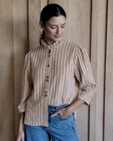 Camisa de corte recto con estampado de rayas verticales en tonos beige y blanco. Presenta cuello alto con detalle de volado, cierre frontal con botones y mangas tres cuartos con sutil fruncido en los hombros.