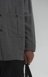 Camisa de manga larga en tejido twill color gris melange, con corte oversize. Presenta cuello clásico, cierre frontal con botones y tres bolsillos delanteros, uno de ellos oculto.