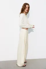Pantalón fluido de color blanco, de tiro alto con trabillas y pliegues en el delantero. Cuenta con bolsillos laterales, cierre frontal con cremallera, botón interior y gancho metálico.