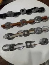 Cinturón marrón de cuero con hebilla plateada rectangular con diseño de flores y apliques metálicos circulares con diseño de flores.