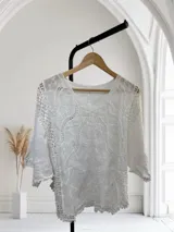 Blusa blanca de manga tres cuartos con cuello cuadrado. Presenta un diseño texturizado con bordados y calados intrincados en el cuerpo y los laterales, con terminación festoneada en el ruedo y puños.
